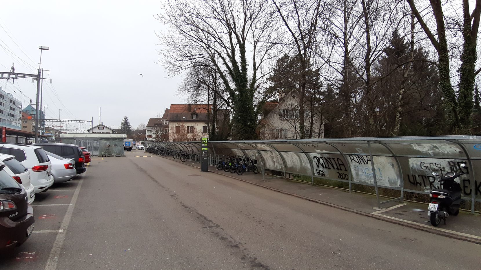 Ein Parkplatz beim Bahnhof Dübendorf mit Autos, daneben ein langer Velounterstand. 